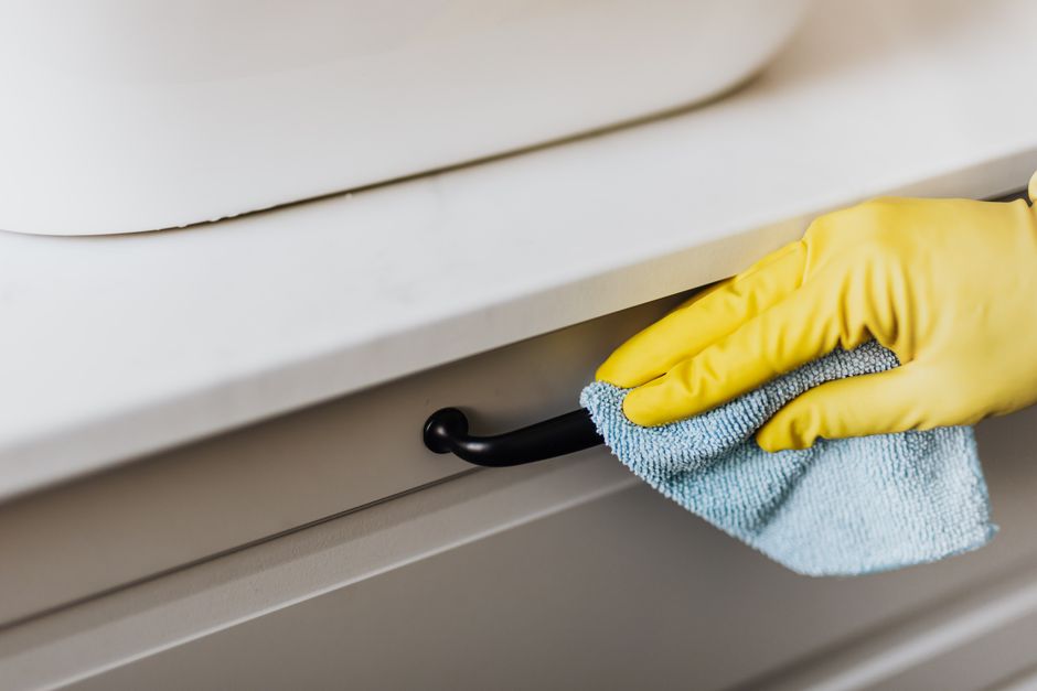 Close-up of a person's hand wearing a yellow rubber glove, wiping the black metal handle of a light grey drawer cabinet in a domestic kitchen setting with a soft blue microfiber cloth. The cabinet sur