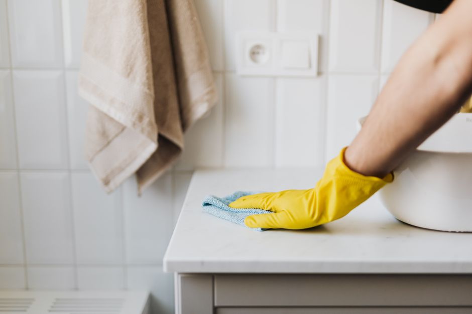 Close-up of a person's hand wearing a yellow rubber glove, wiping the black metal handle of a light grey drawer cabinet in a domestic kitchen setting with a soft blue microfiber cloth. The cabinet sur