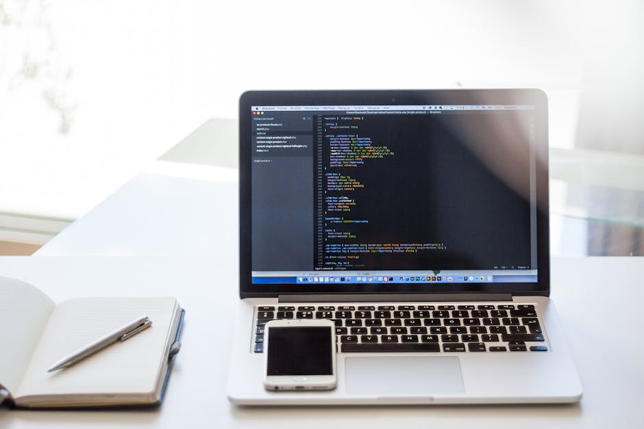 A close-up view of a modern workspace featuring a silver MacBook laptop on a white desk, displaying lines of colorful code on its screen. To the left of the laptop, there is an open blank notebook wit